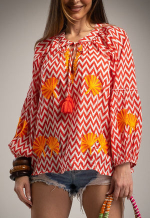 Woman wearing a red and white zigzag patterned top with orange floral designs, holding a woven bag.