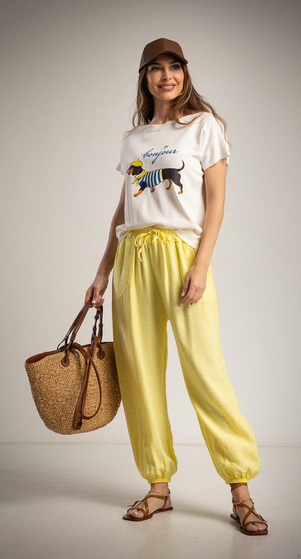 Woman wearing a white t-shirt with a graphic design, yellow pants, and a brown hat, holding a straw bag on a plain background.