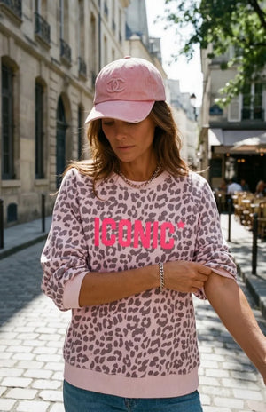 Woman wearing a pink leopard print sweatshirt with 'ICONIC' text and a pink cap in an urban setting.
