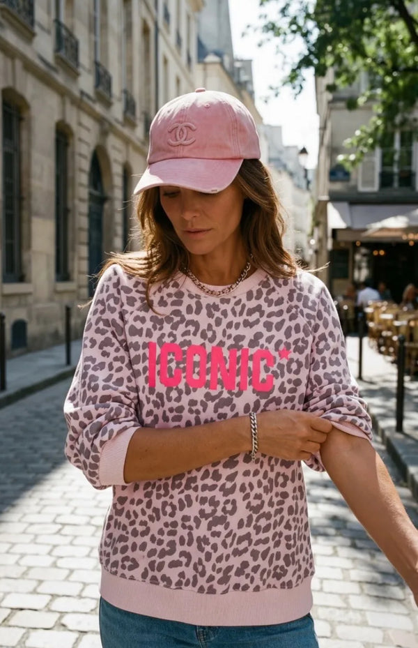 Woman wearing a pink leopard print sweatshirt with 'ICONIC' text and a pink cap in an urban setting.