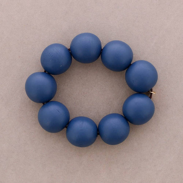 A beaded bracelet made of eight matte blue round beads, arranged in a circle on a neutral taupe background.