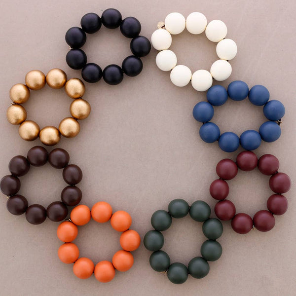 Eight beaded bracelets arranged in a circle on a neutral taupe background, showing different bead colors: black, white, blue, maroon, dark brown, orange, gold, and dark green.