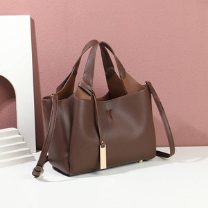 A brown pebbled leather tote bag with dual top handles, a detachable shoulder strap, and a small gold rectangular charm, displayed on a white surface against a pink wall with a white decorative prop.