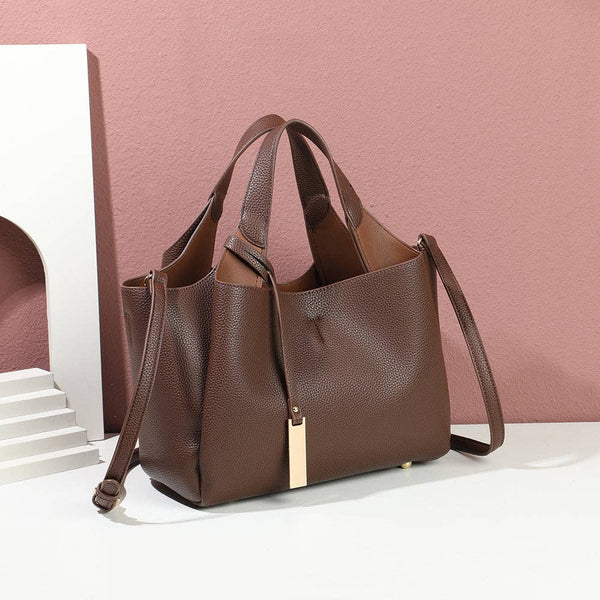 A brown pebbled leather tote bag with dual top handles, a detachable shoulder strap, and a small gold rectangular charm, displayed on a white surface against a pink wall with a white decorative prop.