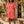 Load image into Gallery viewer, Woman in a red checkered dress standing in a decorative outdoor setting with flowers and stone arches.

