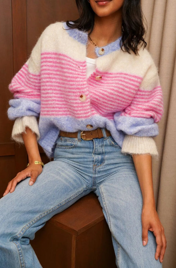 A woman seated indoors wearing a fuzzy knit cardigan with horizontal stripes in cream, pink, and light purple, paired with light blue jeans and a brown belt.