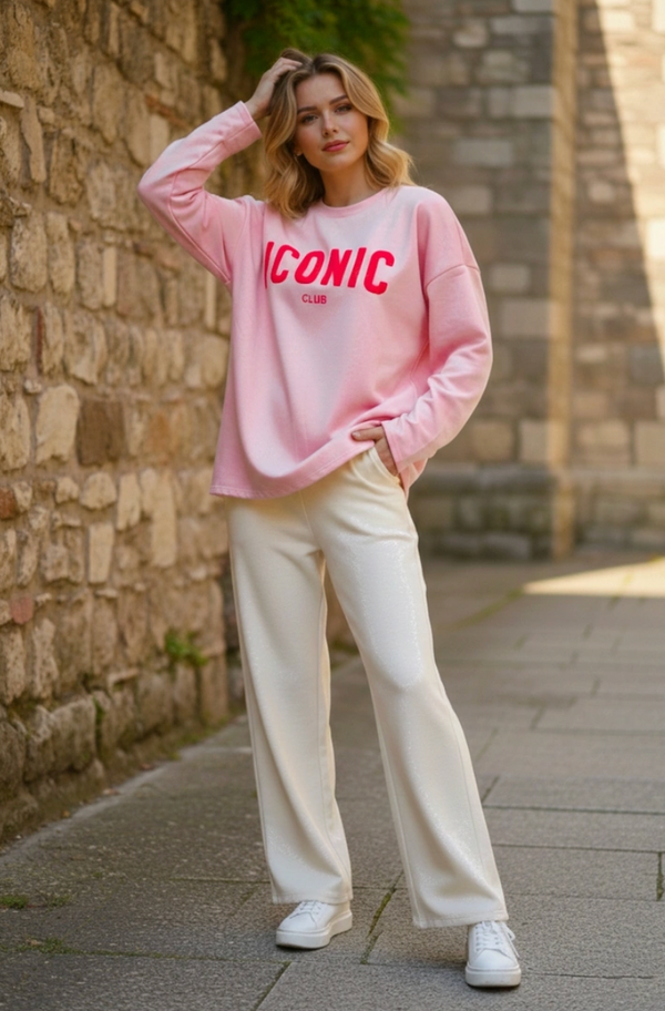 Woman wearing a pink sweatshirt with 'ICONIC' text, white pants, and white sneakers standing against a stone wall.