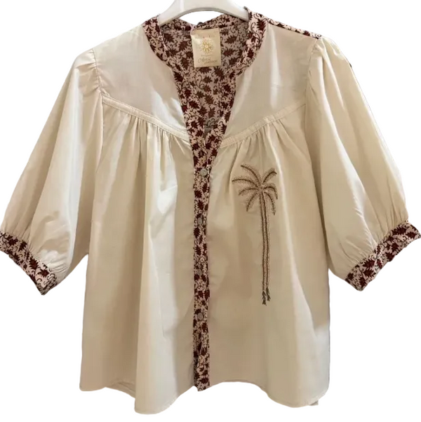 An off-white short-sleeve blouse with a brown and white floral patterned trim, featuring a palm tree embroidery on the chest