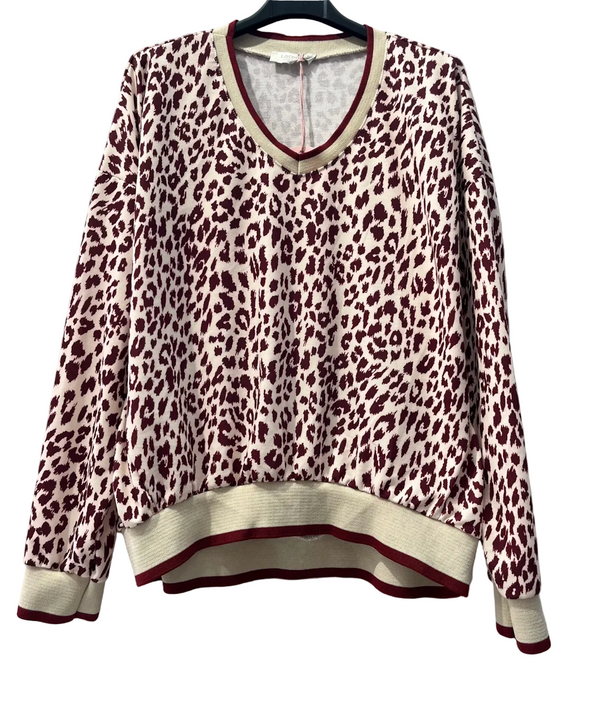 A women's long-sleeve knit sweater with a leopard print pattern in burgundy and cream, featuring ribbed v-neck, cuffs, and hem with matching burgundy and cream trim, hanging on a black hanger against a white brick wall.