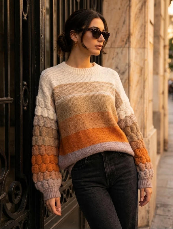 A woman wearing a chunky knit sweater with horizontal stripes in cream, beige, tan, orange, and mauve, paired with dark jeans and sunglasses, leaning against a black wrought-iron gate and beige stone building.