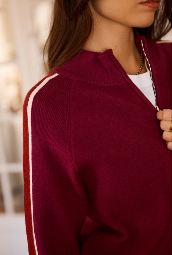Close-up of a woman wearing a burgundy ribbed knit zip-up sweater with a white stripe down the sleeve, over a white top.