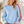 Load image into Gallery viewer, A woman wearing a light blue crewneck sweater with vibrant rainbow stripes down the sleeves, paired with blue jeans, standing in a brightly lit indoor setting.

