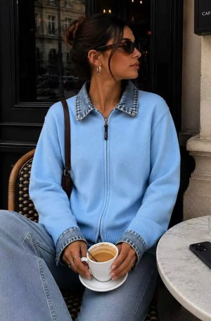 Front view of a woman seated outside a cafe wearing a powder blue fine-knit zip-up cardigan that features a contrast medium-wash denim collar and cuffs, paired with light blue jeans