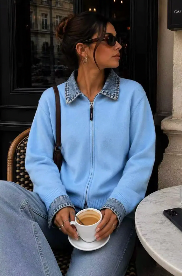 Front view of a woman seated outside a cafe wearing a powder blue fine-knit zip-up cardigan that features a contrast medium-wash denim collar and cuffs, paired with light blue jeans