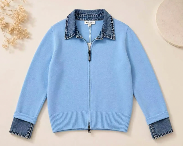 A light blue zip-up knit cardigan with a denim collar, hem, and cuffed sleeves, laid flat on a light beige background with dried flowers and a circular prop.