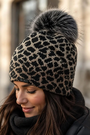 "Woman wearing a black and beige leopard print knit beanie hat with a large grey faux fur pom pom out in the cold" 