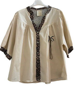 A cream-colored short-sleeve blouse with leopard print trim along the V-neckline, button placket, and cuffs, featuring a small black palm tree applique on the chest, hanging on a white hanger against a textured concrete wall.