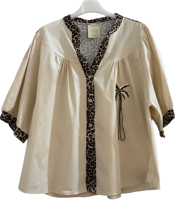 A cream-colored short-sleeve blouse with leopard print trim along the V-neckline, button placket, and cuffs, featuring a small black palm tree applique on the chest, hanging on a white hanger against a textured concrete wall.