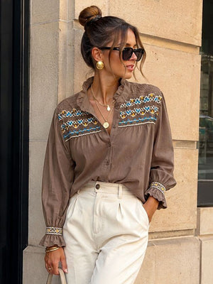 A woman stands on a city sidewalk wearing a brown long-sleeve blouse with colorful geometric embroidery across the chest, paired with high-waisted white wide-leg trousers, beige pointed-toe heels, and carrying a beige handbag.