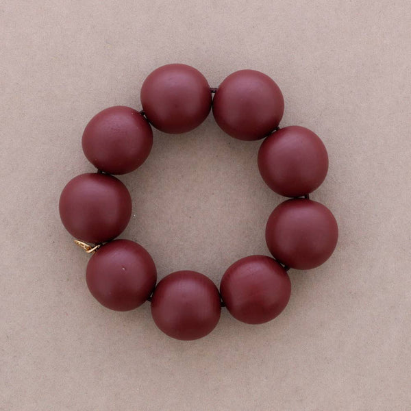 A beaded bracelet made of matte, dark red-brown round beads arranged in a circle on a neutral beige background.