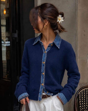 Front view of a woman wearing a navy blue fine-knit button-up cardigan featuring a contrast light-wash denim collar, placket, and layered cuffs, finished with polished gold dome buttons and styled with white pants