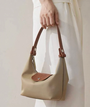 Beige handbag with brown leather accents held by a person wearing a white dress.